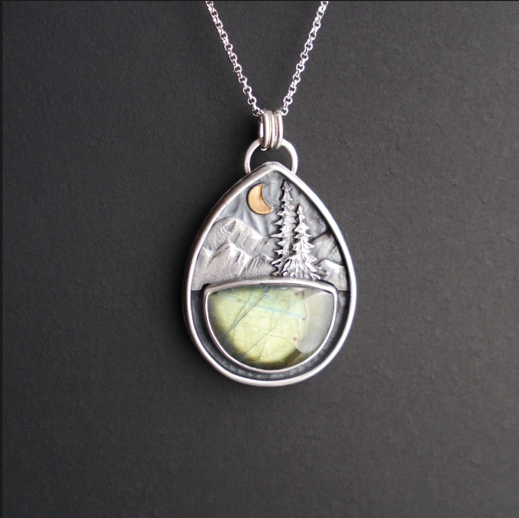 Teardrop shaped silver pendant hanging from a silver chain with a sea green labradorite half moon stone beneath textured mountains and evergreen trees with a gold crescent moon. 