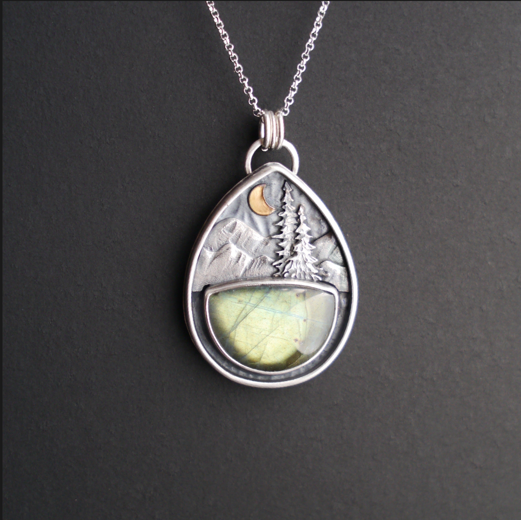 Teardrop shaped silver pendant hanging from a silver chain with a sea green labradorite half moon stone beneath textured mountains and evergreen trees with a gold crescent moon.